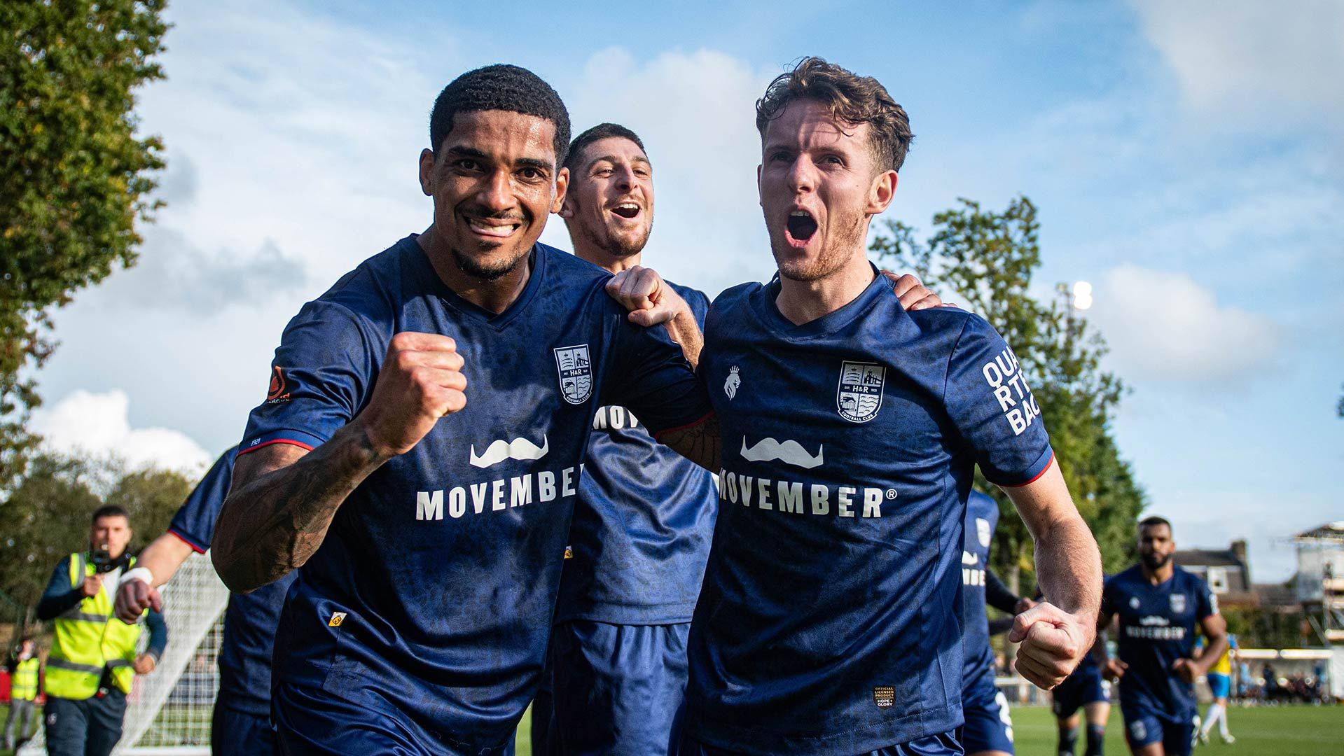 Photo showing three men at a football field, celebrating and wearing Movember-branded football attire.